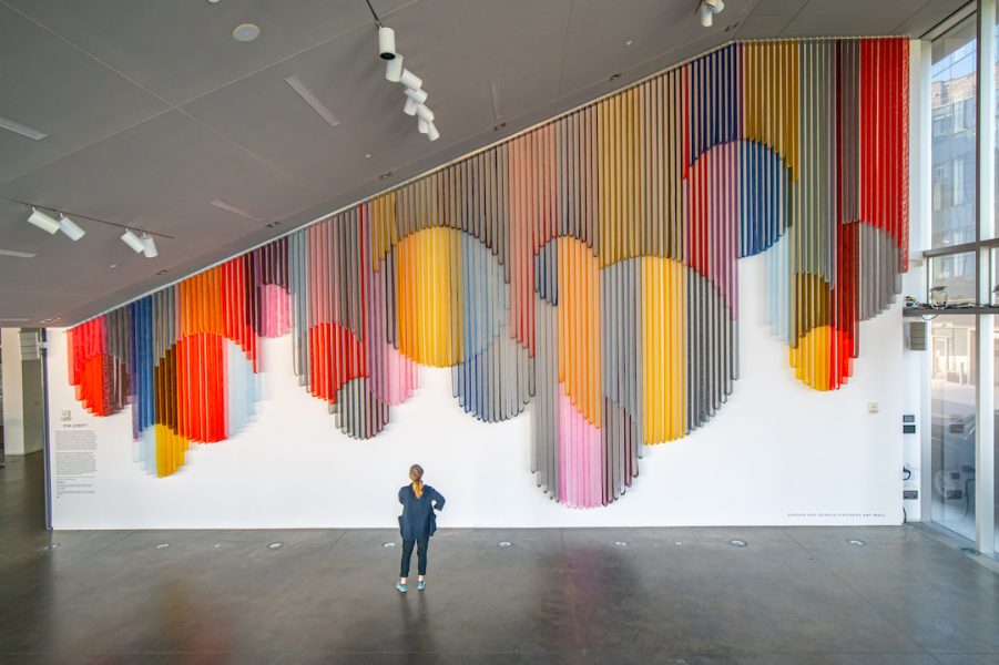 A visitor standing in an empty spacious lobby and looking up at a monumental hanging wall sculpture made from bands of colorful coated mesh fabric which drape in various lengths to create series of interlocking circular forms. 