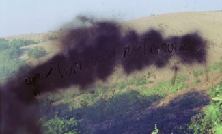 A color photograph of a sandy dune with plants obscured by a cloudy, black form with the words "yes Tomorrow no Tomorrow" scratched into it.