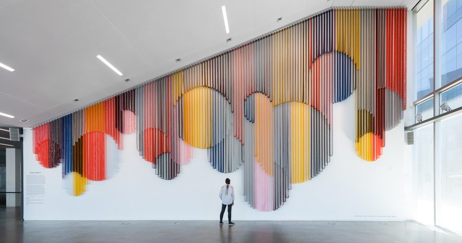 A visitor standing in a spacious lobby and looking up at a monumental hanging wall sculpture made from bands of colorful coated mesh fabric draped in various lengths to create series of interlocking circular forms.