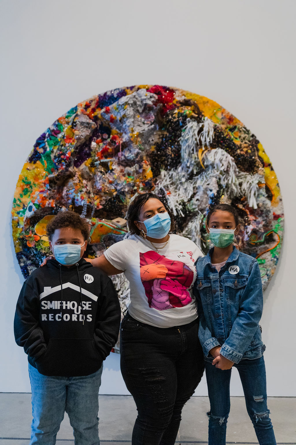 A mother and her two children pose for a photo in front of a large round, multicolored and textured collage