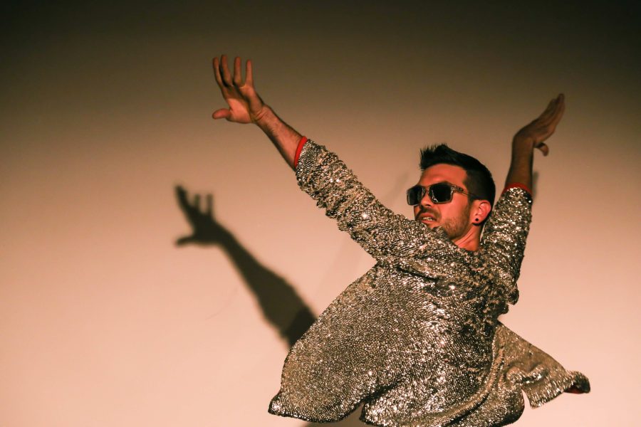 A man wearing sunglasses and a glittery silver jacket strikes a pose with arms outstretched.