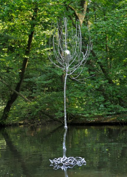 A stainless steel sculpture of a bare tree, with a human head depicted at the top of the trunk, suspended in a body of water in a forest.
