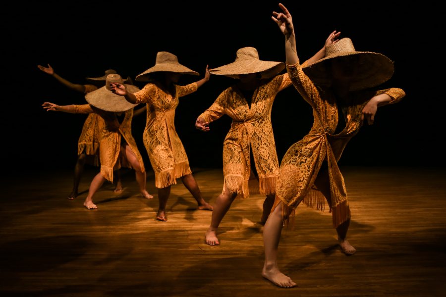 A line of performers wearing wide straw hats and sheer lace robes in dramatic stage lighting