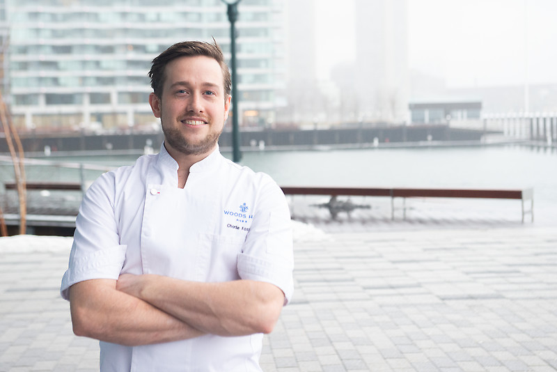 Chef Charlie Foster, in his chef's uniform, arms crossed and smiling, with Boston Harbor in the background.
