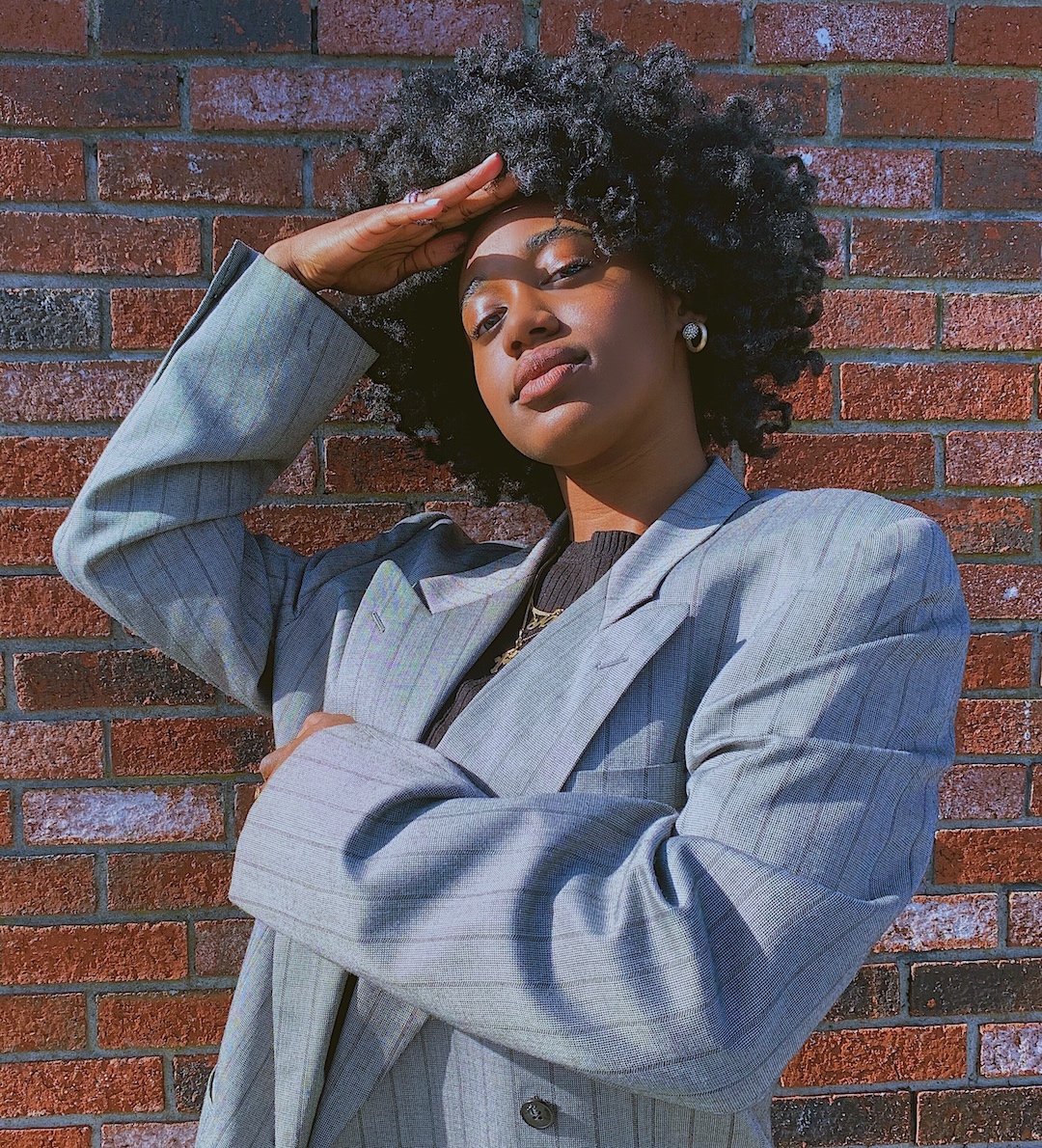 A dark skinned woman posing and wearing a suit jacket in front a brick wall.