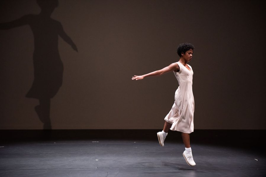 A dancer in a pale dress hops with arms stretched behind her on a dark stage.