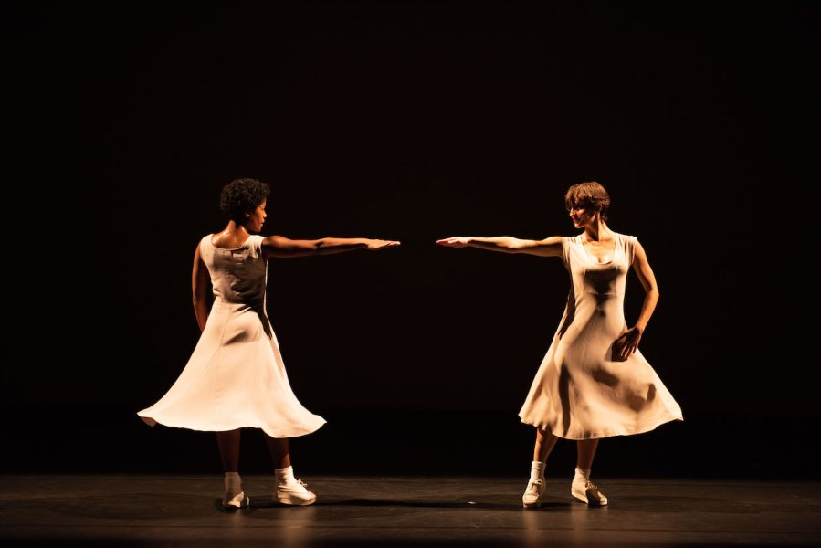 Two female dancers in pale dresses stand on a dark stage with one arm extended toward the other.
