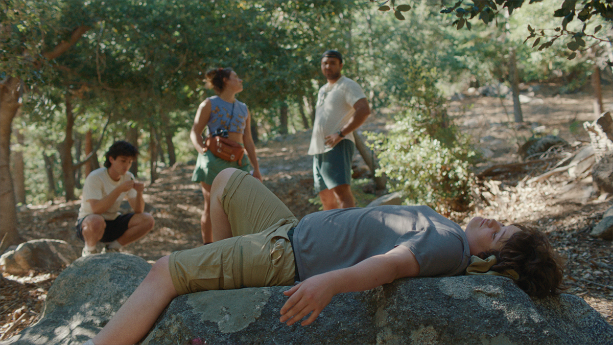 A boy lying down on some rocks outside with eyes closed with a few people standing in the distance