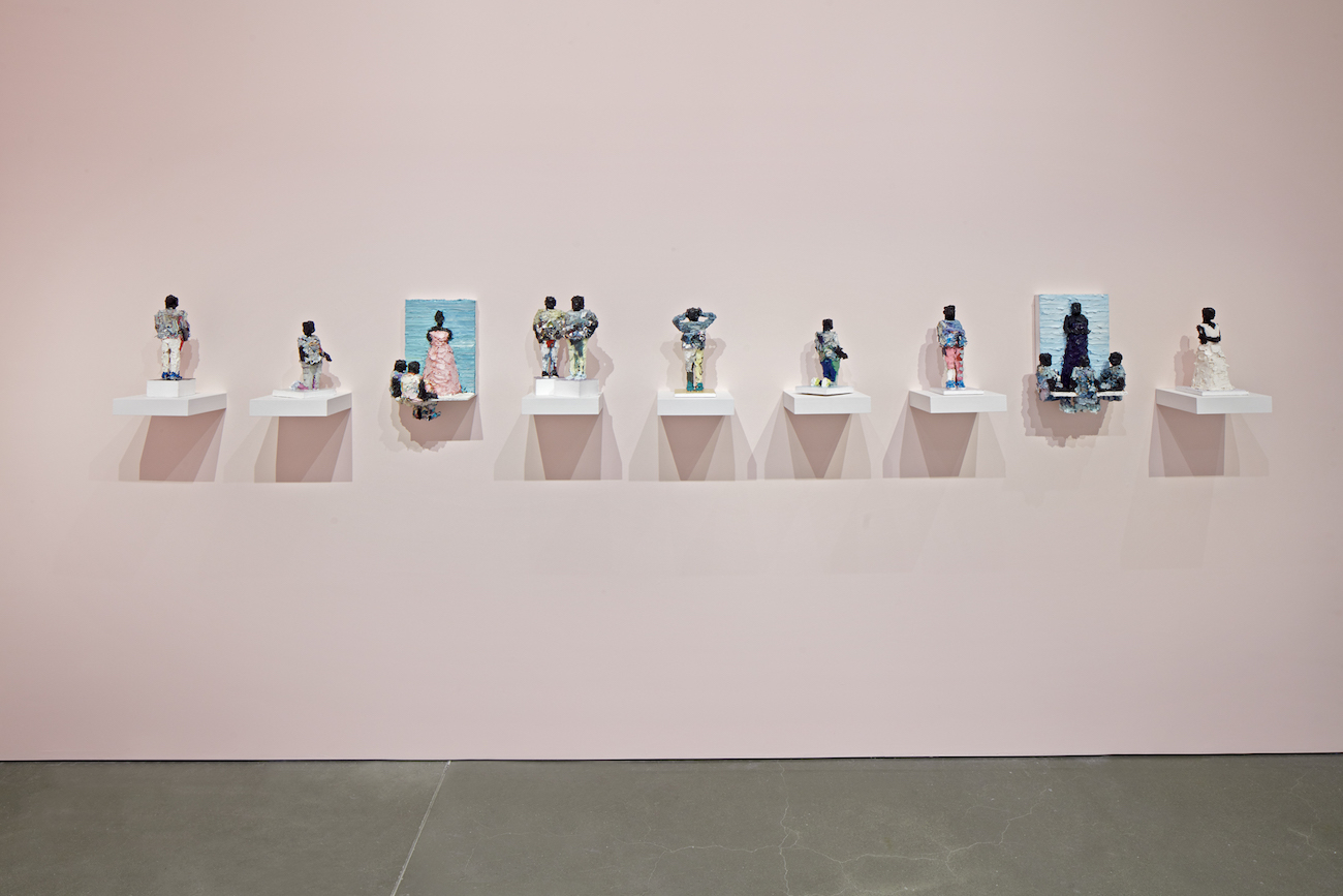 A pale pink wall features 9 small figures made out of paint sitting on small white wall-mounted pedestals.