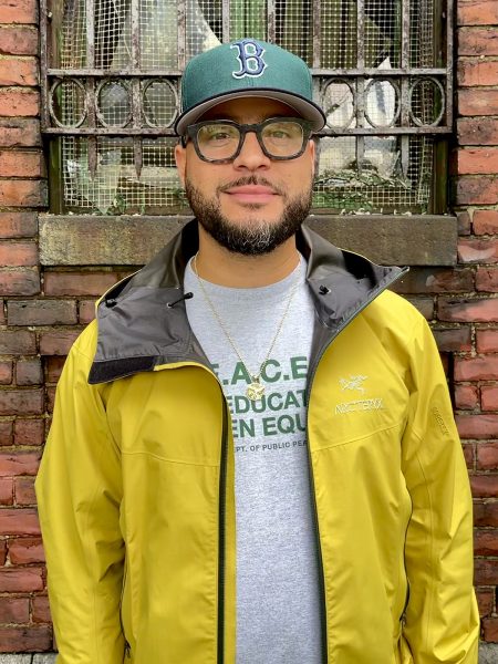 Image of man with Snapback and yellow jacket standing against a brick wall