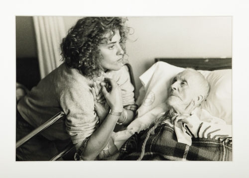 A black-and-white photograph of a light-skinned young woman leaning over the railing of a hospital bed, holding the hand of a frail and pale elderly woman who lays in the bed and looks up at her. 