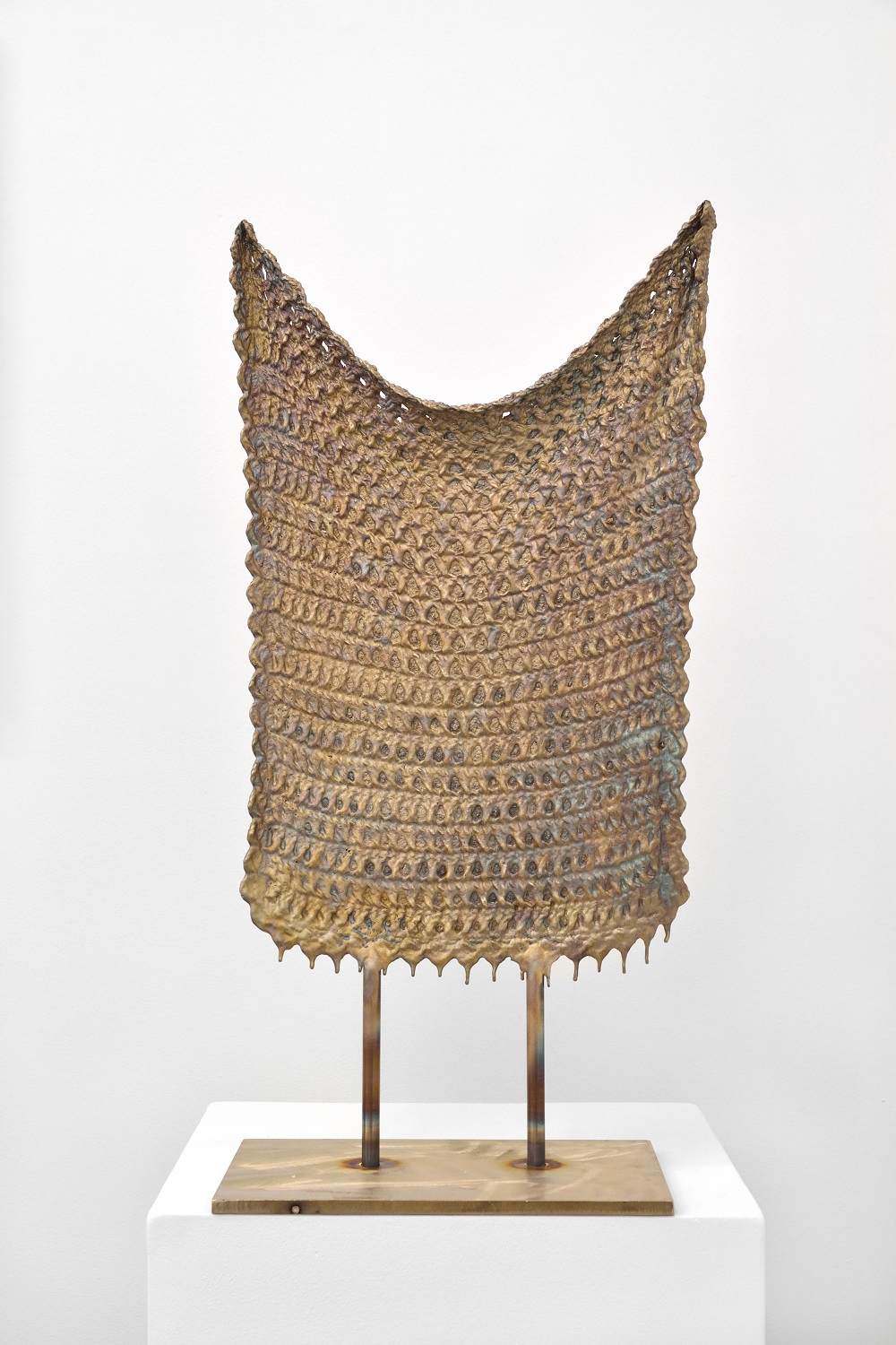 A bronze sculpture of a knitted blanket suspended between two rods on a narrow metal plinth.