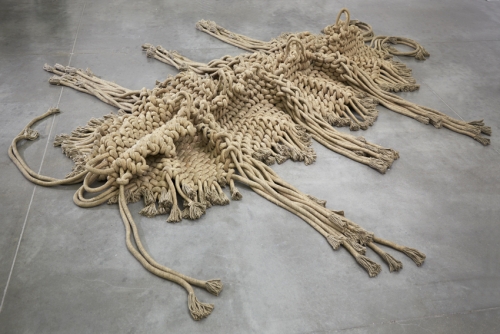A sculpture made of very thick, beige rope or cordwoven together to resemble an abstracted inchworm on a concrete floor.