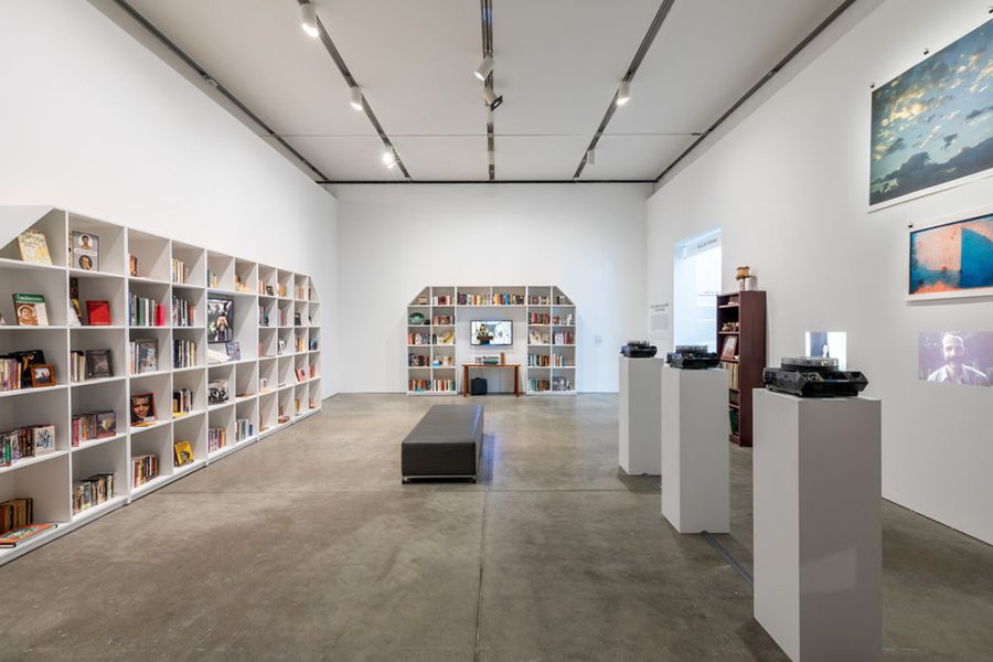 Gallery with large bookshelves filled with books and assorted objects, as well as projected and hanging images along the right wall and a bench in the middle of the room.