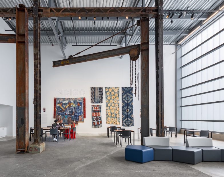 A room with white walls, iron beams and gantries, a selection of colorful fabric wall hangings under the title 