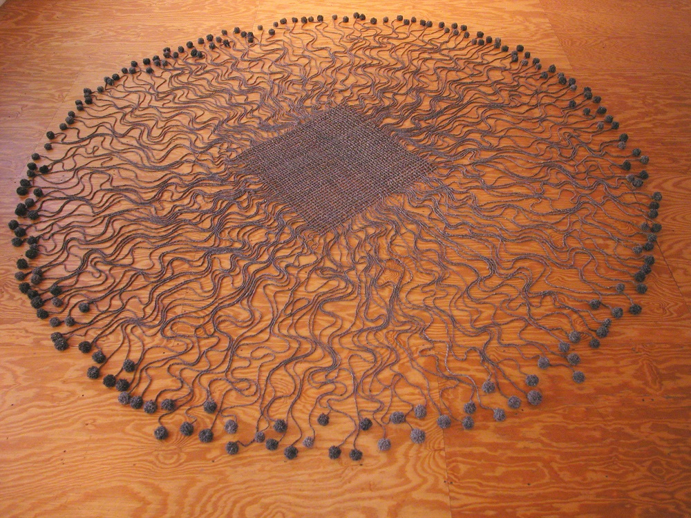 A floor sculpture of gray natural wool consisting of a woven square with individual threads radiating in wavy lines and ending in miniature balls of yarn to form a loose circle enclosing the square.