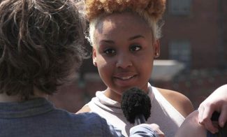 A video still shows a medium-dark-skinned young woman speaking into a microphone and looking at her interviewer, whose back is to the viewer.
