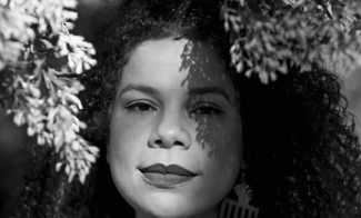 A black-and-white portrait of artist Firelei Báez, a light skinned Latinx woman with curly hair standing underneath a tree.