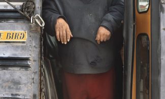 A color photograph depicts a full portrait of a light-skinned man in dirty, ragged clothing standing in the open doors of a school bus as he looks directly at the viewer.