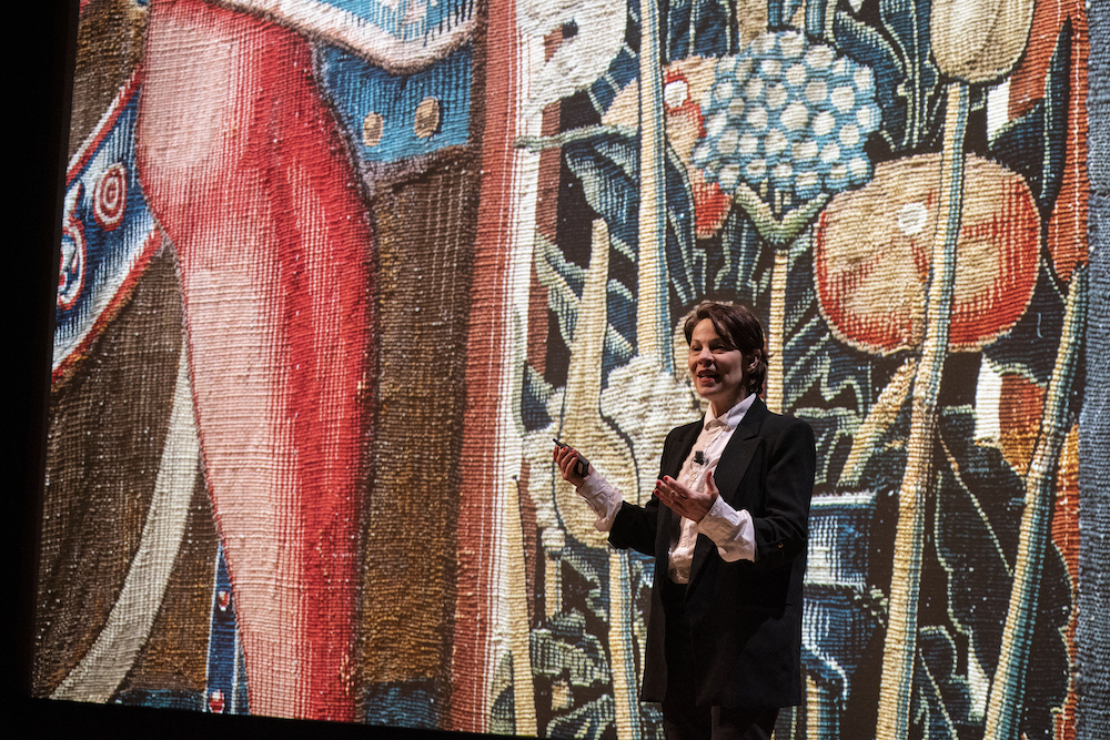 A woman standing in front of a tapestry lecturing. 
