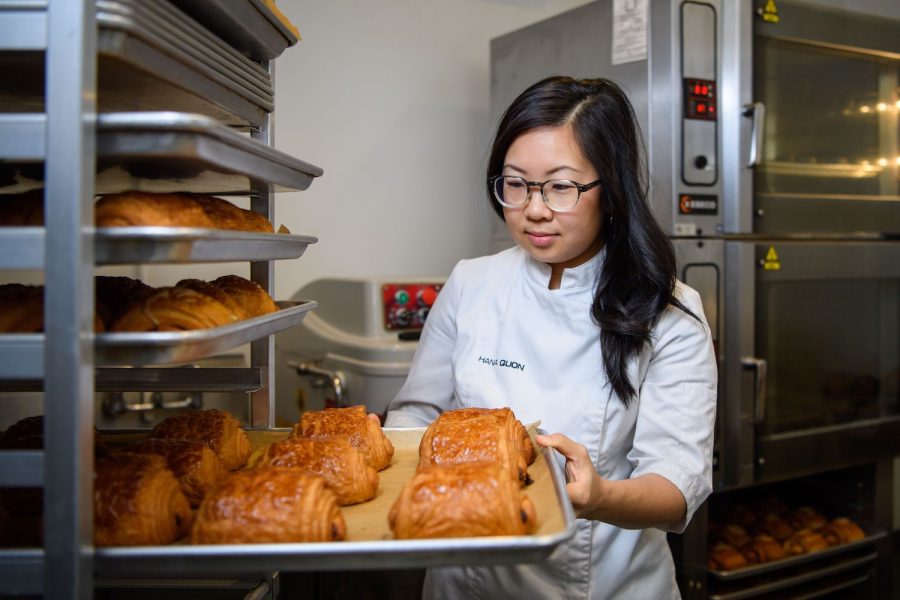 A chef with brown skin, glasses, and log black hair taking a tray of pastries off of a shelf in an industrial kitchen.