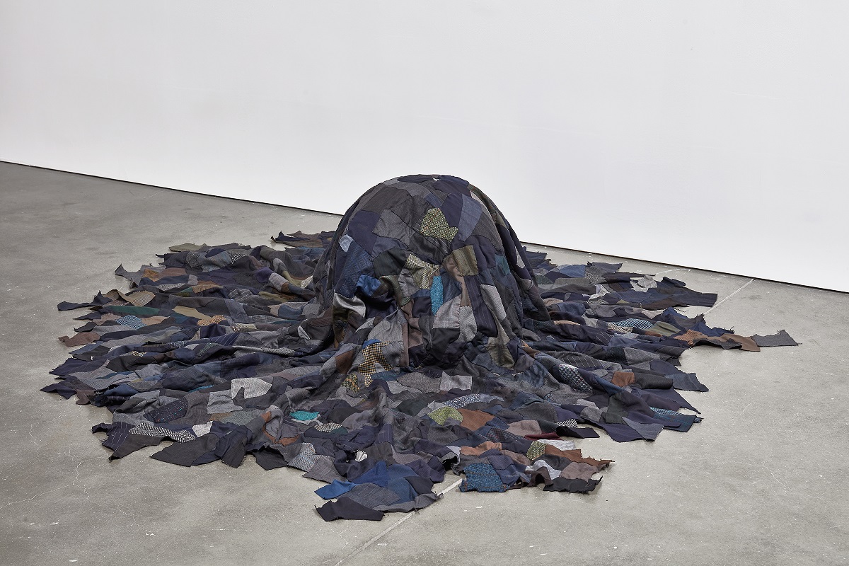 A sculpture of stitched fabric squares in dark shades over a mound reminiscent of a crouched body on the floor of an empty gallery.