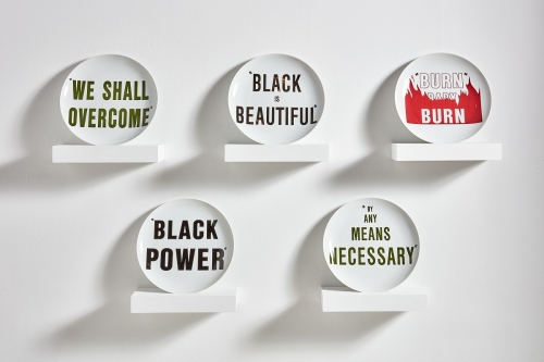 An installation of five ceramic plates displayed on five shelves and printed with the sayings