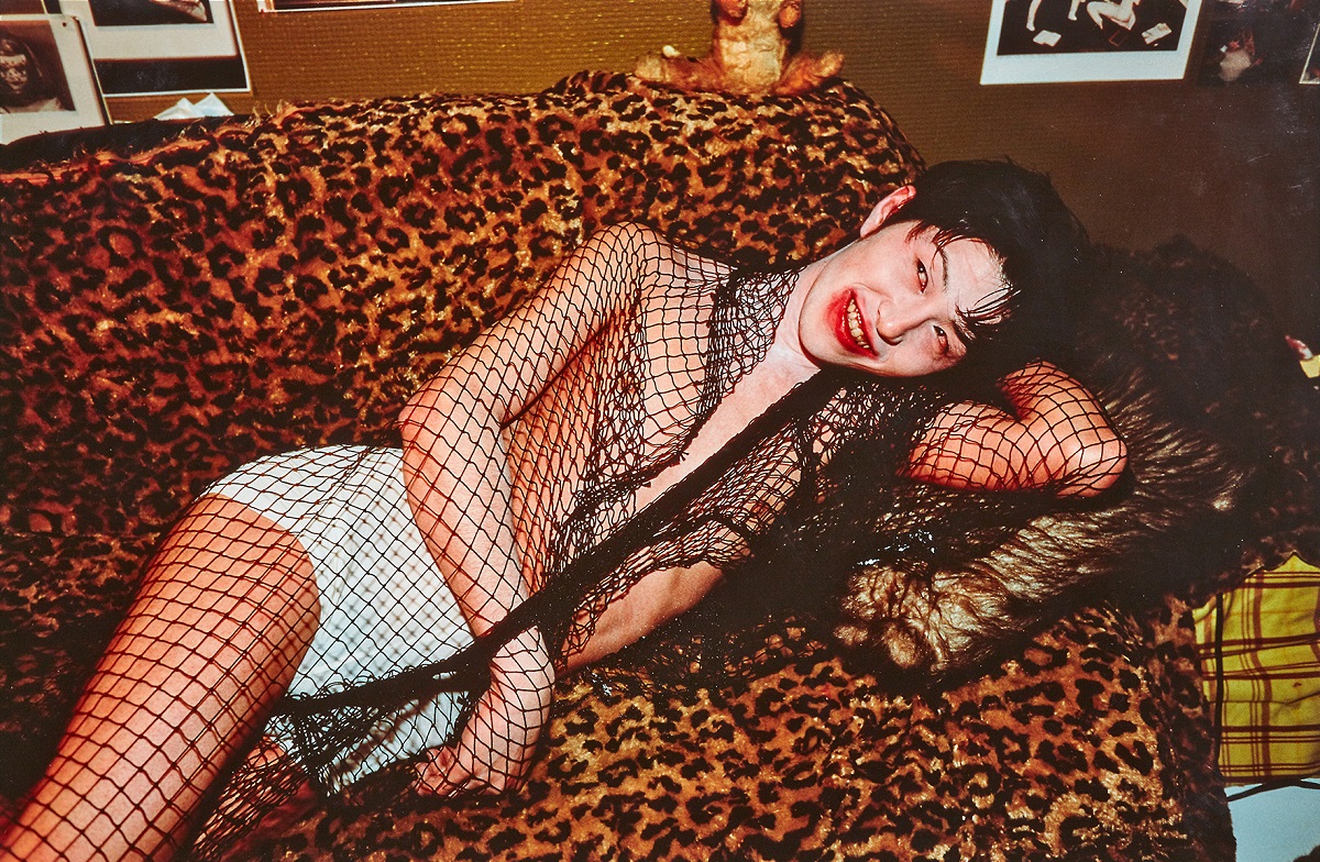 A color photograph of a Japanese, male-presenting young adult laying on their side on a leopard print sofa, smiling through smudged red lipstick, and wearing a black fishnet scarf over white underpants.