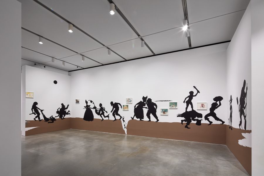 A photograph of an installation comprising black, life-size silhouettes on a brown ground acting out a series of scenes across three walls of a gallery.