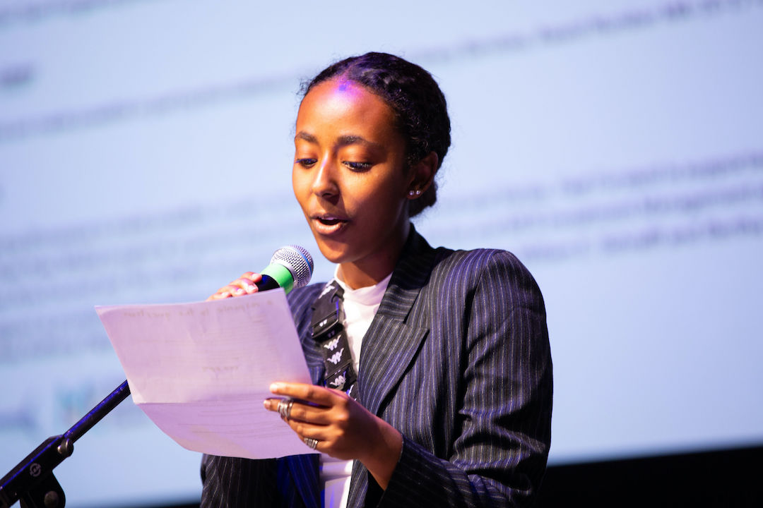 A medium-dark skinned young person speaks into a mic while reading off a paper with a monitor projection behind them.