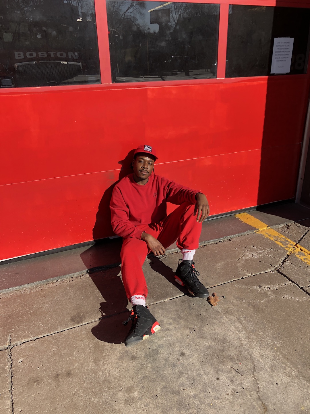 A man wearing all red sitting on a pavement against a red-colored building.