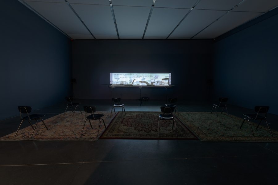 A dark space with a narrow-framed moving images projection illuminating the gallery filled with school chairs and three rugs.