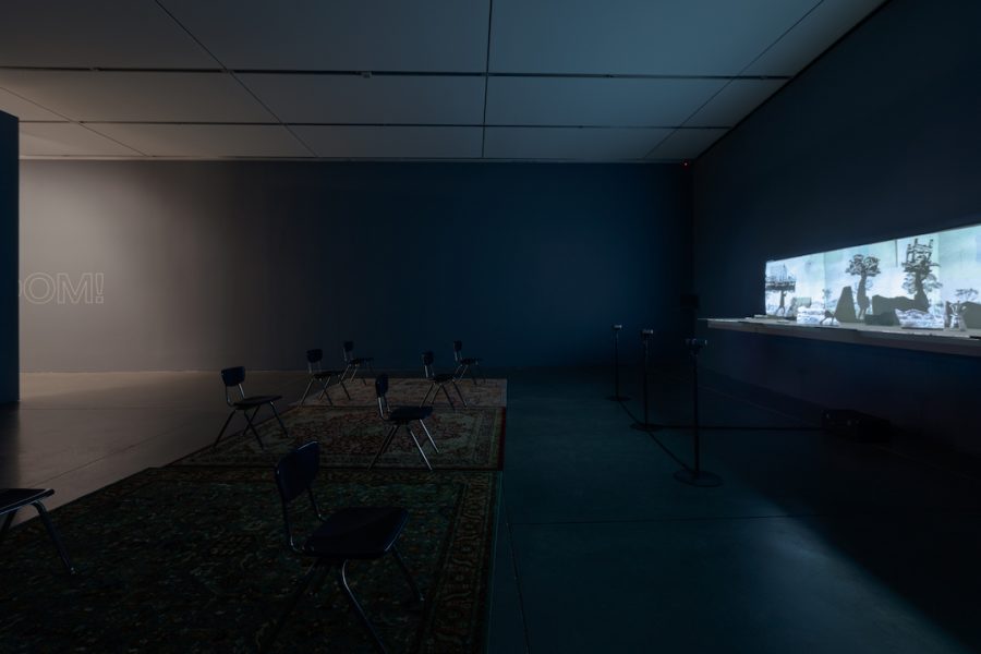 A side angle of a dark space with a moving image projection illuminating the gallery filled with school chairs and three rugs.