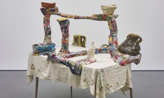 A sculpture consisting of a colorful ceramic and papier mache structure with multiple arms holding ceramic vessels and displayed on a table with an embroidered white tablecloth.