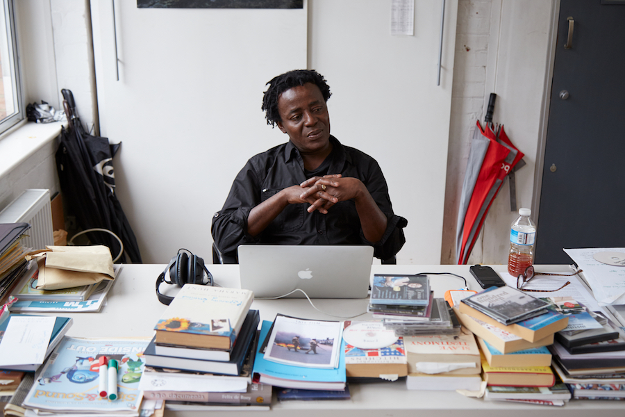 British-Ghanian artist John Akomfrah sits with folded hands behind a white desk stacked with books and CDs. He looks down and to his left and is wearing a black button-up-shirt with the sleeves pushed up.