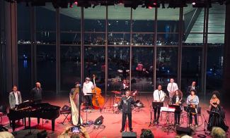 An array of musicians and instruments on the ICA's stage at night while a man speaks into the microphone