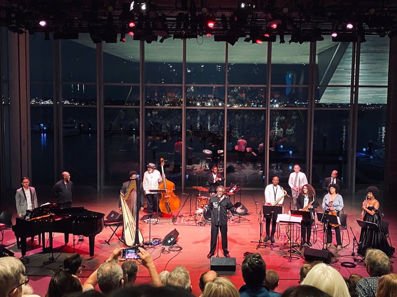An array of musicians and instruments on the ICA's stage at night while a man speaks into the microphone