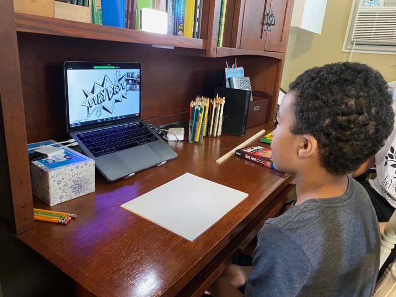 Young Black boy sat at a desk facing a piece of paper and laptop in front of him. 