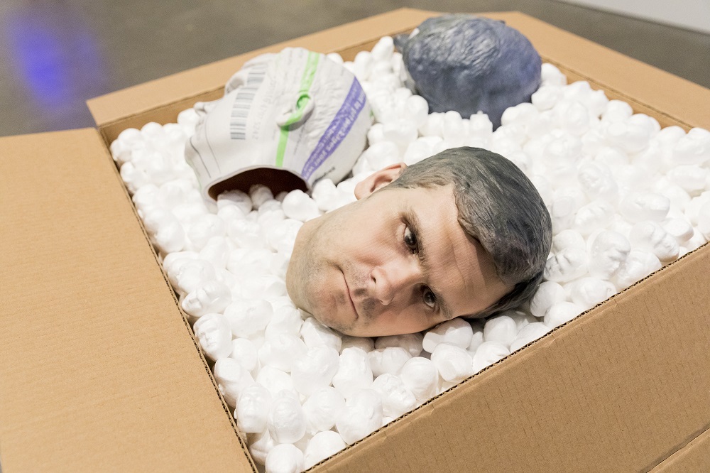 A sculpture shows the 3D-printed head of a light-skinned man lying on its side in an open cardboard box filled with white packing peanuts, beside two other hollowed heads of indeterminate material.
