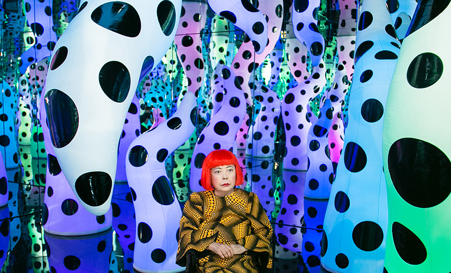 Yayoi Kusama, with bright red hair and a yellow patterned shirt, sits in her Infinity Mirror Room, LOVE IS CALLING, a mirrored room filled with inflated glowing tentacles in different colors and colored with black dots of varying sizes. 