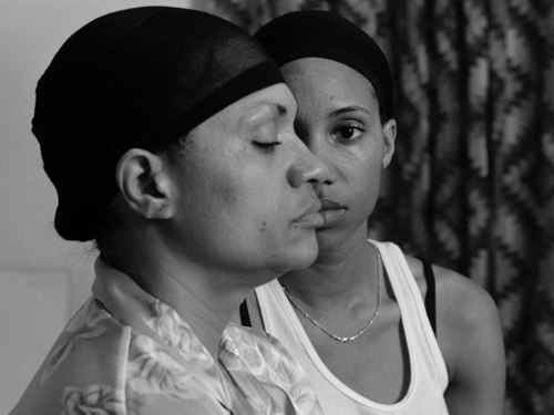 A black-and-white photograph shows the artist, a Black woman, looking directly at the viewer as she stands behind her mother, who is shown in profile and whose head obscures half of the artist's face. Both are wearing hair caps and a curtain is seen in the background.