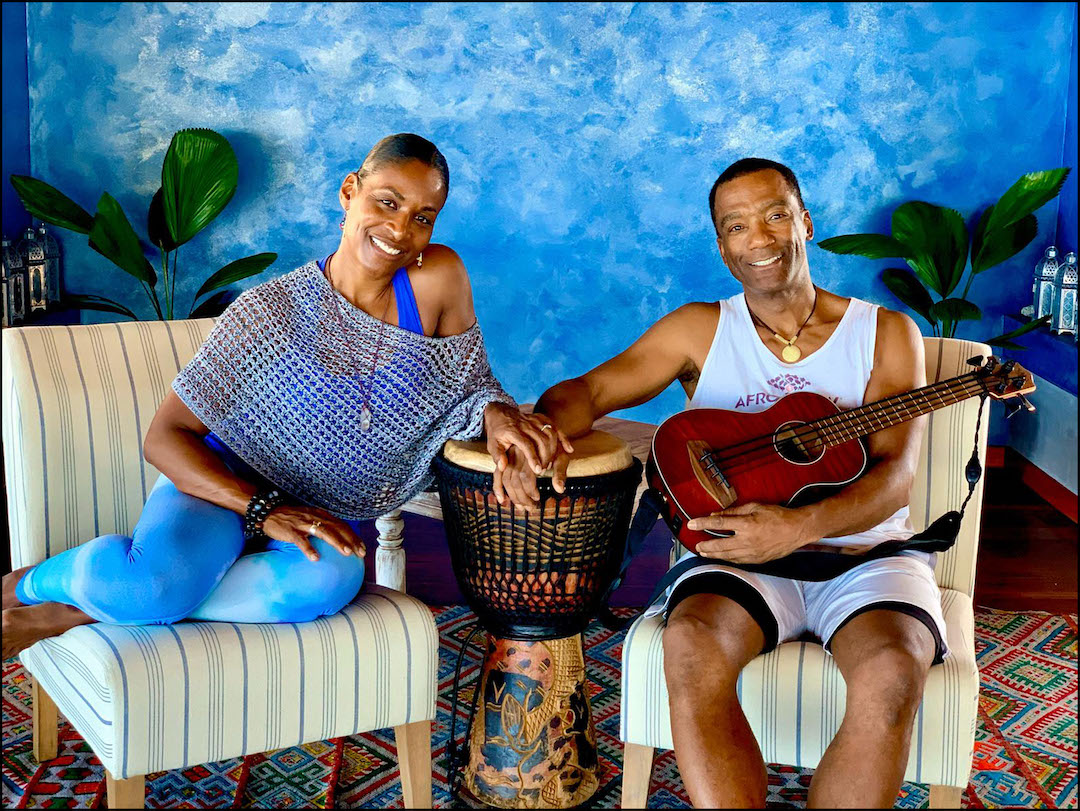 Two people, a wife and husband, sitting in a tropical interior room with their hands touching over a djembe drum and the husband holding a guitar.