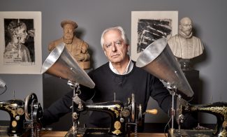 A portrait of artist William Kentridge, an elderly white man, standing behind a table with antique sewing machines and silver megaphones attached to them; behind him are classical bust sculptures and prints.