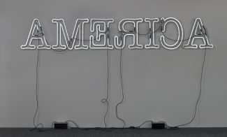 A white neon sculpture hung on a wall spells "AMERICA" in backwards capital letters.