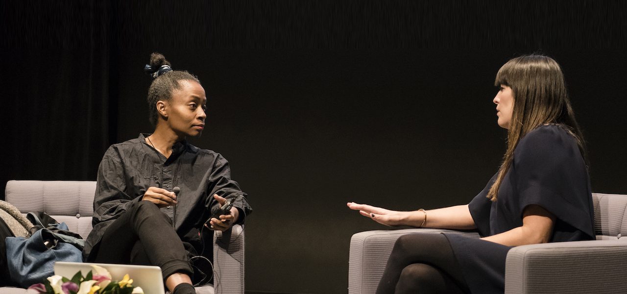 The artist Kara Walker and curator Eva Respini sit and talk onstage in gray chairs against a black background