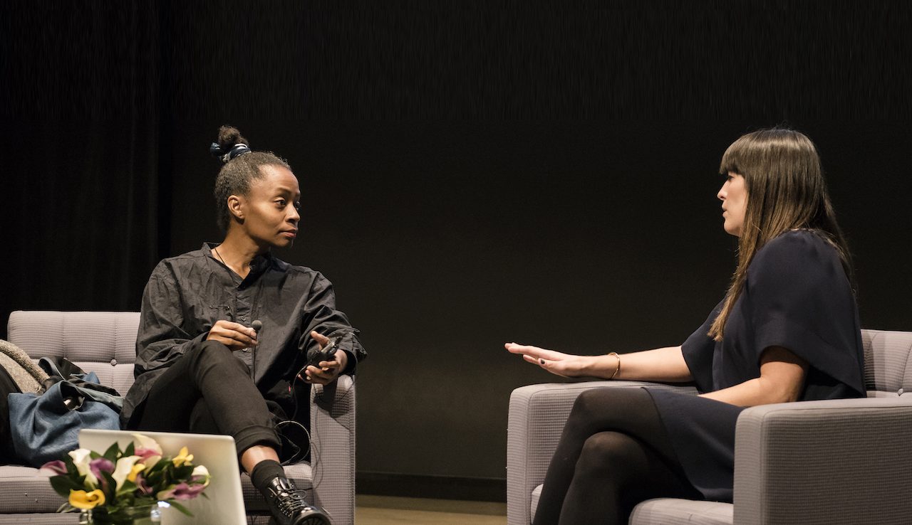 The artist Kara Walker and curator Eva Respini sit and talk onstage in gray chairs against a black background