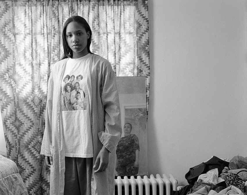 A black-and-white photograph shows the artist, a Black woman, gazing directly at the viewer with the reflection of her mother visible in a mirror behind her. She is wearing a T-shirt with the cast of the Cosby Show on it.