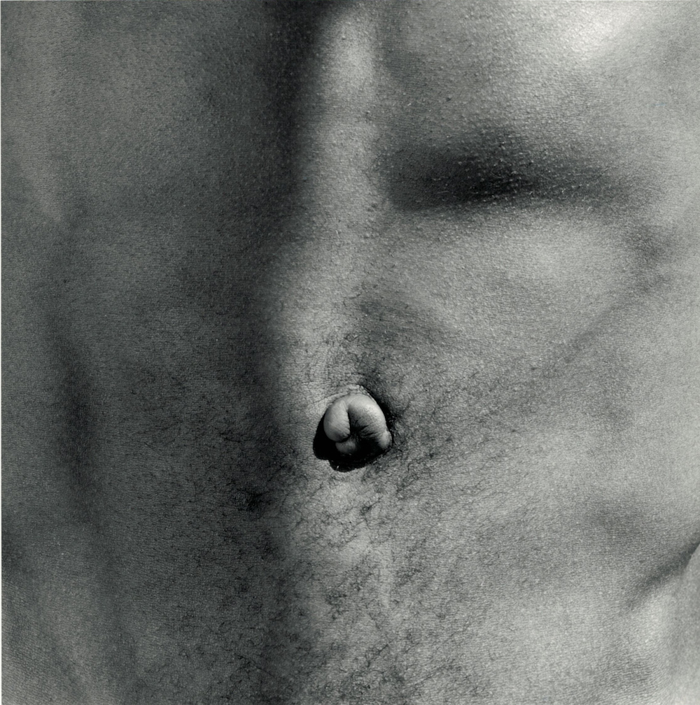 A black-and-white square photograph shows a tight close up of a light-skinned man’s flat, bare stomach with a belly button in the center.