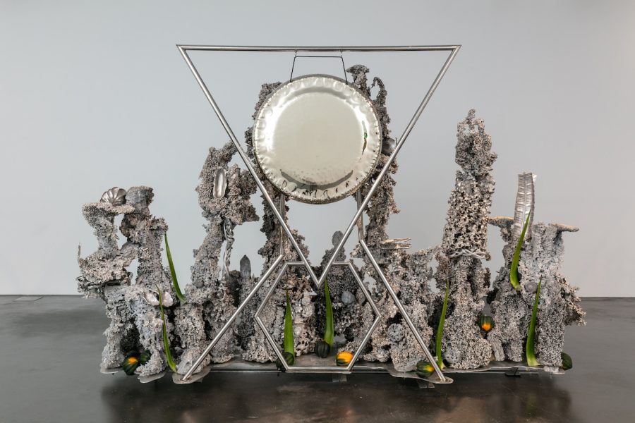 A silvery mixed-media sculpture with a hanging cymbal-like form surrounded by a number of standing, indistinct forms.