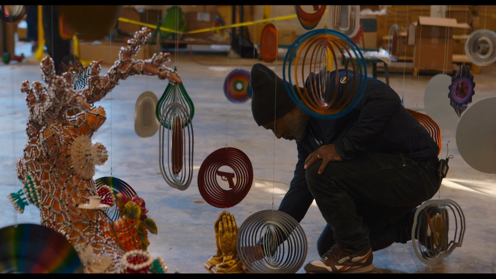 Film still shows artist Nick Cave, in a dark knit hat and clothing, crouching down among multiple hanging decorative reflectors.